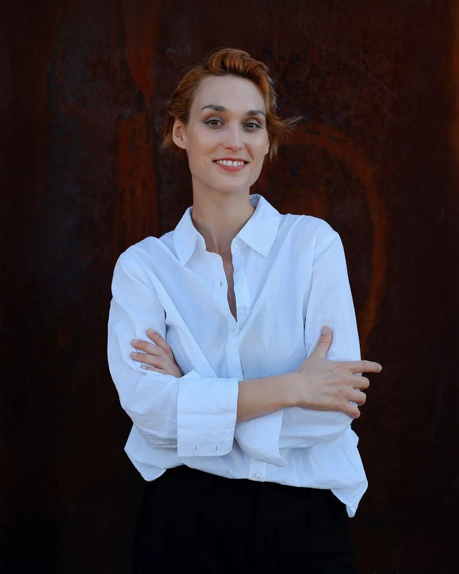 Professional headshot of actor Deborah Muriel Blum in a crisp white shirt, confidently posed against a deep red textured wall