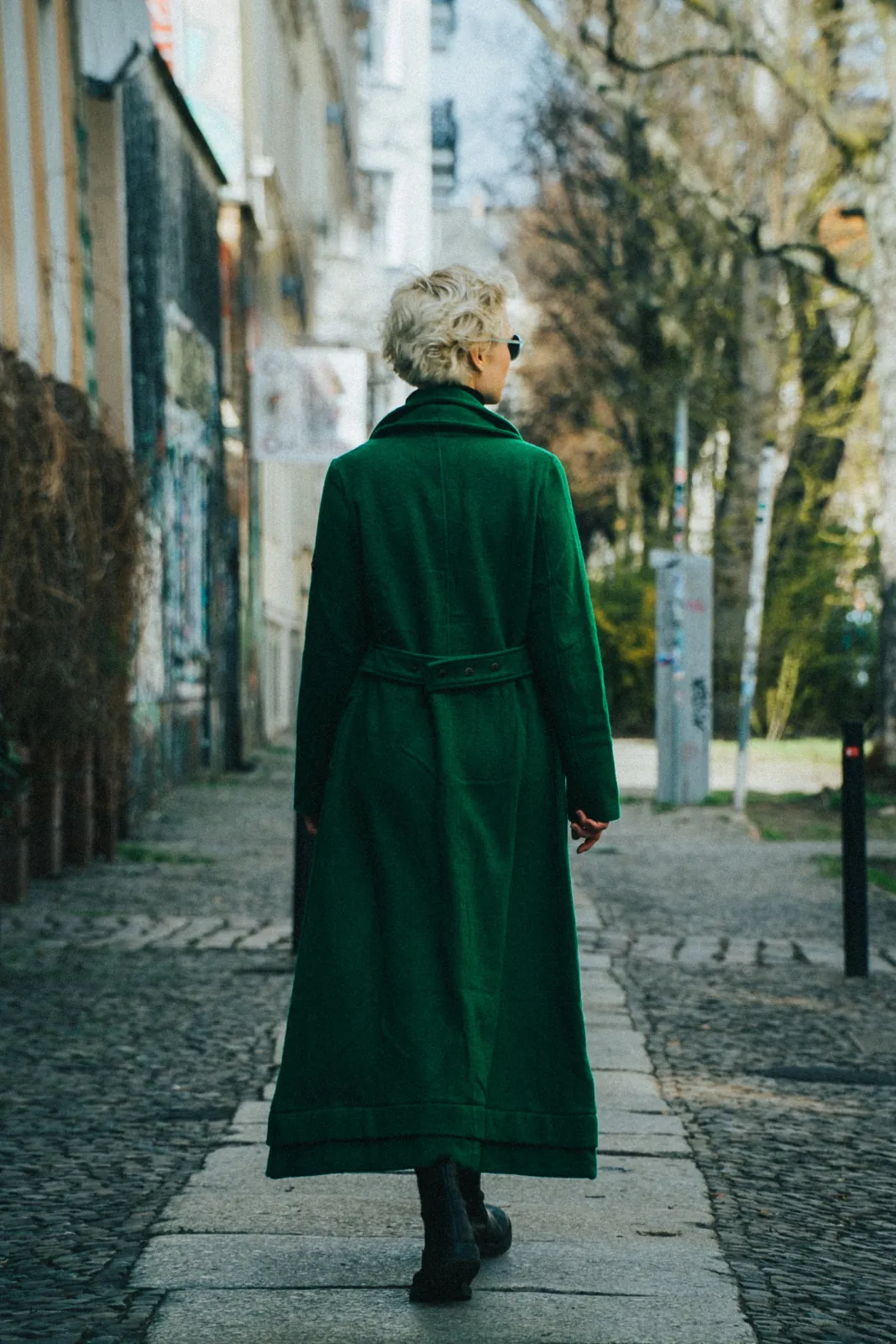 Deborah Muriel Blum walking a Berlin street in a long green coat